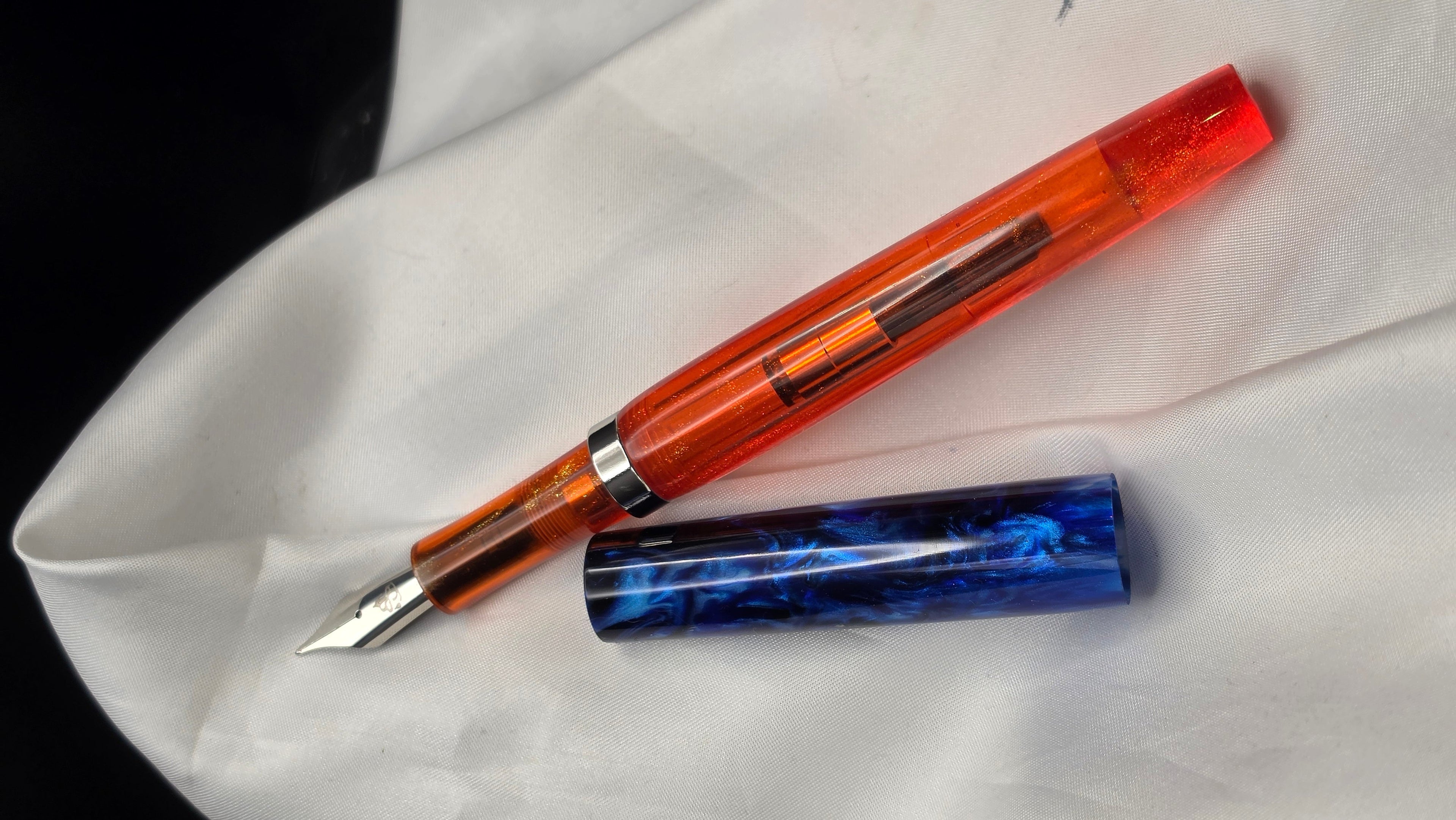 A translucent orange fountain pen with a silver nib, shown next to its blue marbled cap on a draped white fabric background.